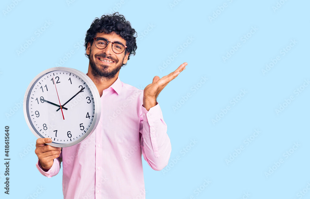 Handsome young man with curly hair and bear holding big clock celebrating victory with happy smile and winner expression with raised hands Handsome young man with curly hair and bear holding big clock celebrating victory with happy smile and winner expression with raised hands