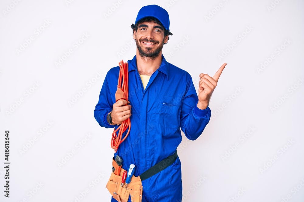 Handsome young man with curly hair and bear wearing builder jumpsuit uniform and electric cables smiling happy pointing with hand and finger to the side
