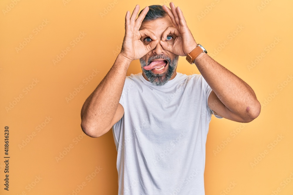 Middle age handsome man wearing casual white tshirt doing ok gesture like binoculars sticking tongue out, eyes looking through fingers. crazy expression. Middle age handsome man wearing casual white tshirt doing ok gesture like binoculars sticking tongue out, eyes looking through fingers. crazy expression.