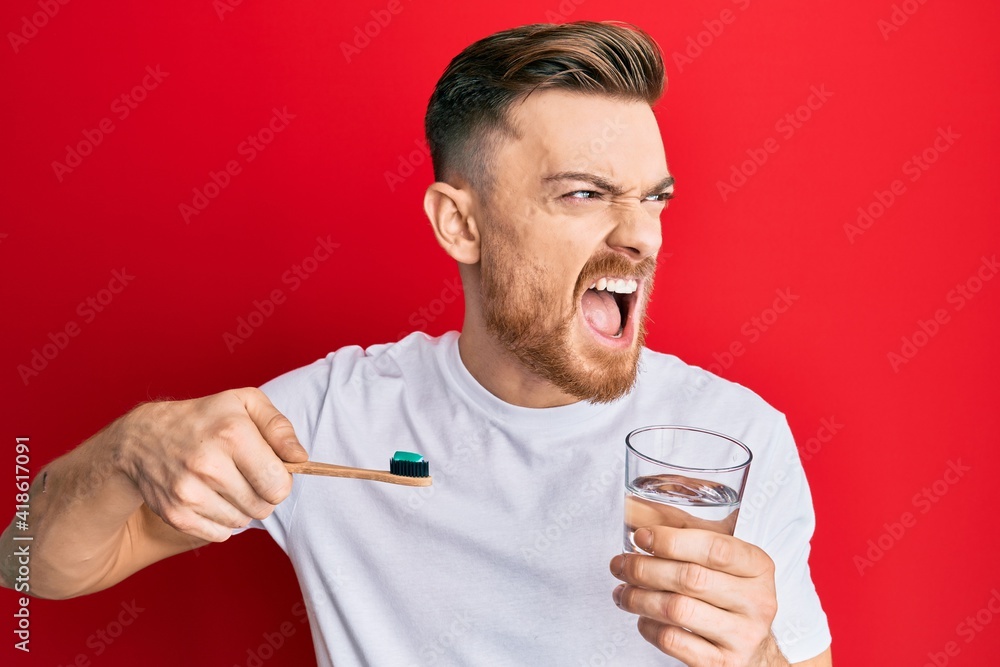 Young redhead man holding toothbrush with toothpaste and glass of water angry and mad screaming frustrated and furious, shouting with anger. rage and aggressive concept. Young redhead man holding toothbrush with toothpaste and glass of water angry and mad screaming frustrated and furious, shouting with anger. rage and aggressive concept.