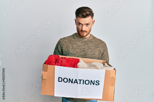 Young redhead man volunteer holding donations box skeptic and nervous, frowning upset because of problem. negative person.