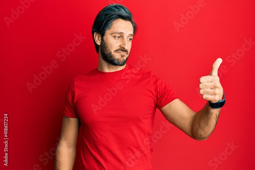 Young hispanic man wearing casual red t shirt looking proud, smiling doing thumbs up gesture to the side