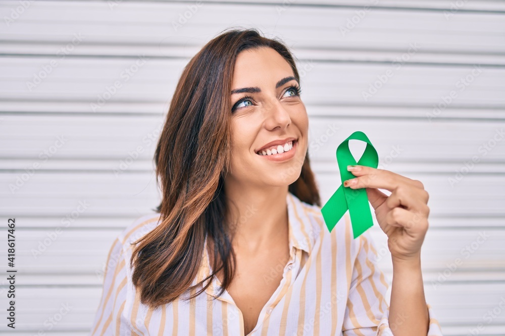 Young hispanic woman smiling happy holding awareness green ribbon standing at the city. Young hispanic woman smiling happy holding awareness green ribbon standing at the city.