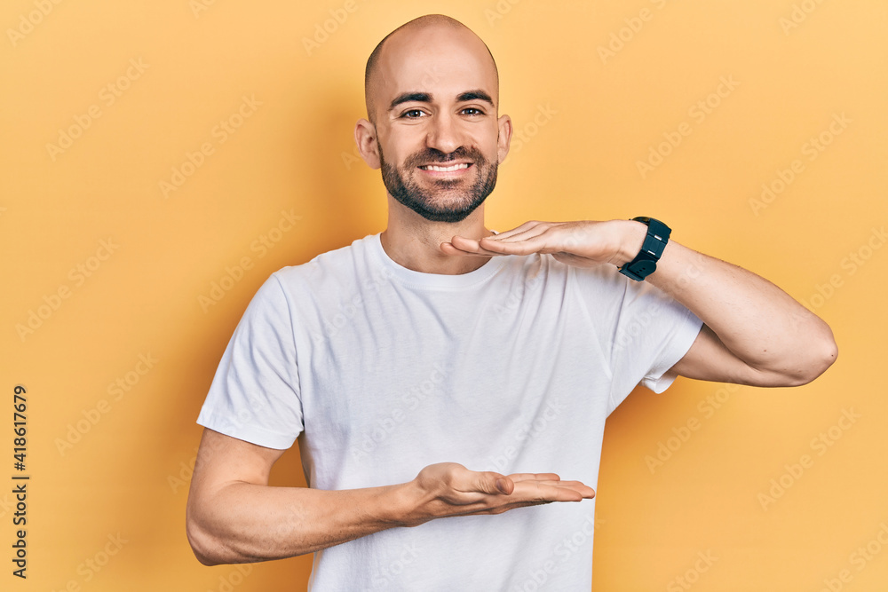 Young bald man wearing casual white t shirt gesturing with hands showing big and large size sign, measure symbol. smiling looking at the camera. measuring concept. Young bald man wearing casual white t shirt gesturing with hands showing big and large size sign, measure symbol. smiling looking at the camera. measuring concept.
