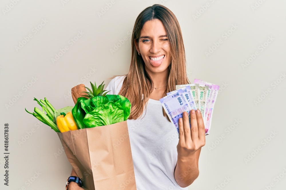 Brunette young woman holding groceries and indian rupee banknotes sticking tongue out happy with funny expression. Brunette young woman holding groceries and indian rupee banknotes sticking tongue out happy with funny expression.