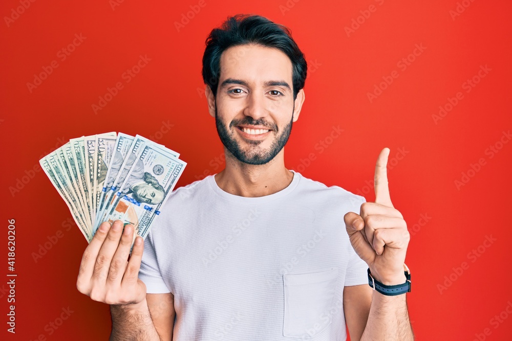 Young hispanic man holding dollars smiling with an idea or question pointing finger with happy face, number one Young hispanic man holding dollars smiling with an idea or question pointing finger with happy face, number one