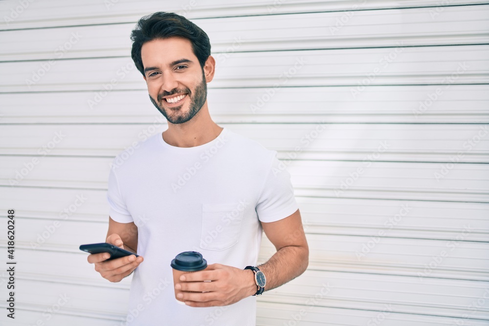 Fototapeta premium Young hispanic man smiling happy using smartphone and drinking take away coffee at city.
