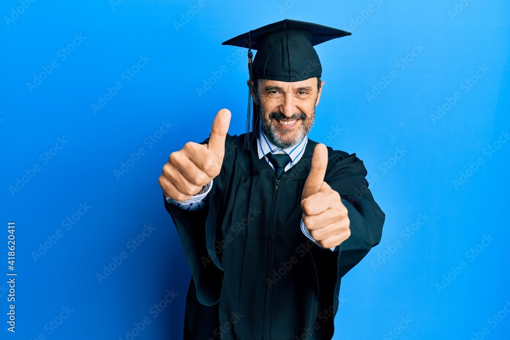 Middle age hispanic man wearing graduation cap and ceremony robe approving doing positive gesture with hand, thumbs up smiling and happy for success. winner gesture. Middle age hispanic man wearing graduation cap and ceremony robe approving doing positive gesture with hand, thumbs up smiling and happy for success. winner gesture.