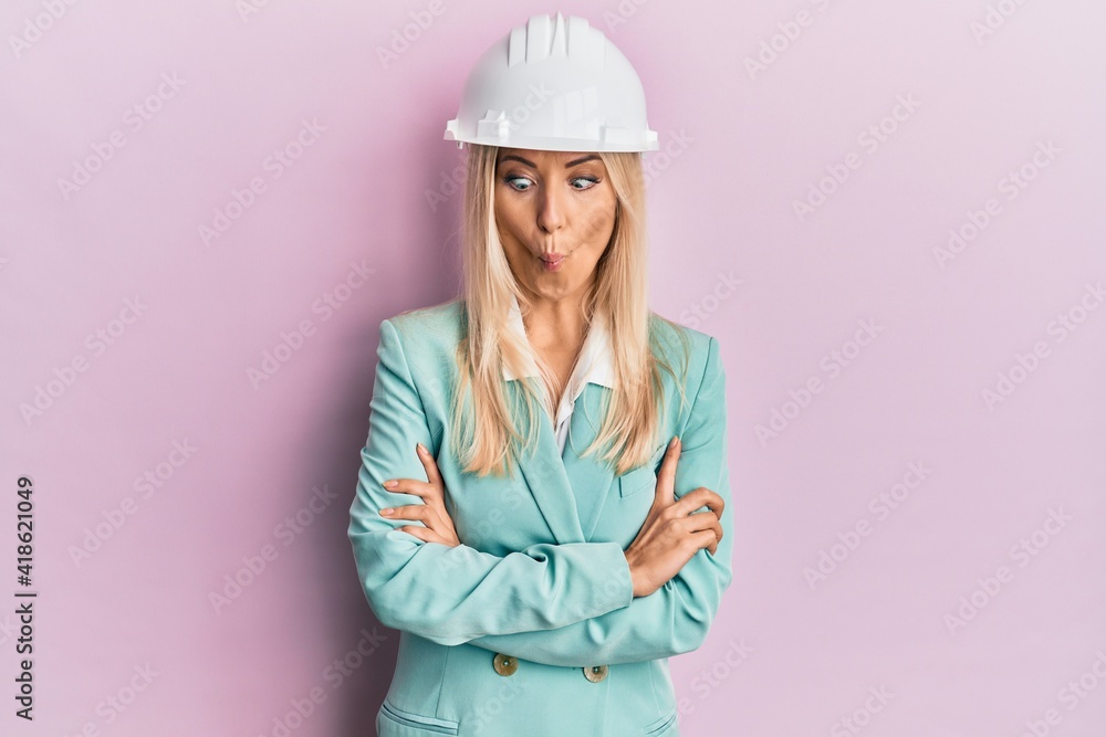 Young blonde woman wearing architect hardhat making fish face with mouth and squinting eyes, crazy and comical.