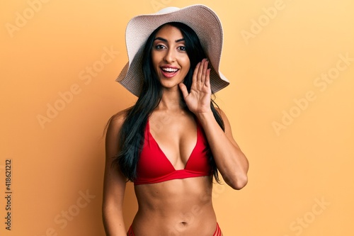 Beautiful hispanic woman wearing bikini and summer hat smiling with hand over ear listening and hearing to rumor or gossip. deafness concept.