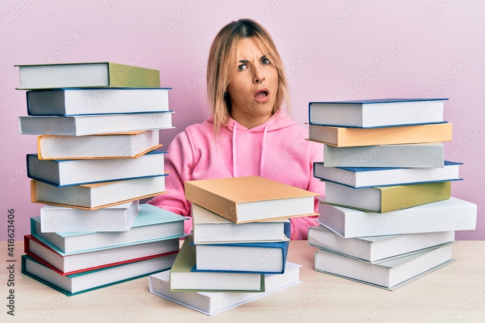 Young caucasian woman sitting on the table with books in shock face, looking skeptical and sarcastic, surprised with open mouth Young caucasian woman sitting on the table with books in shock face, looking skeptical and sarcastic, surprised with open mouth