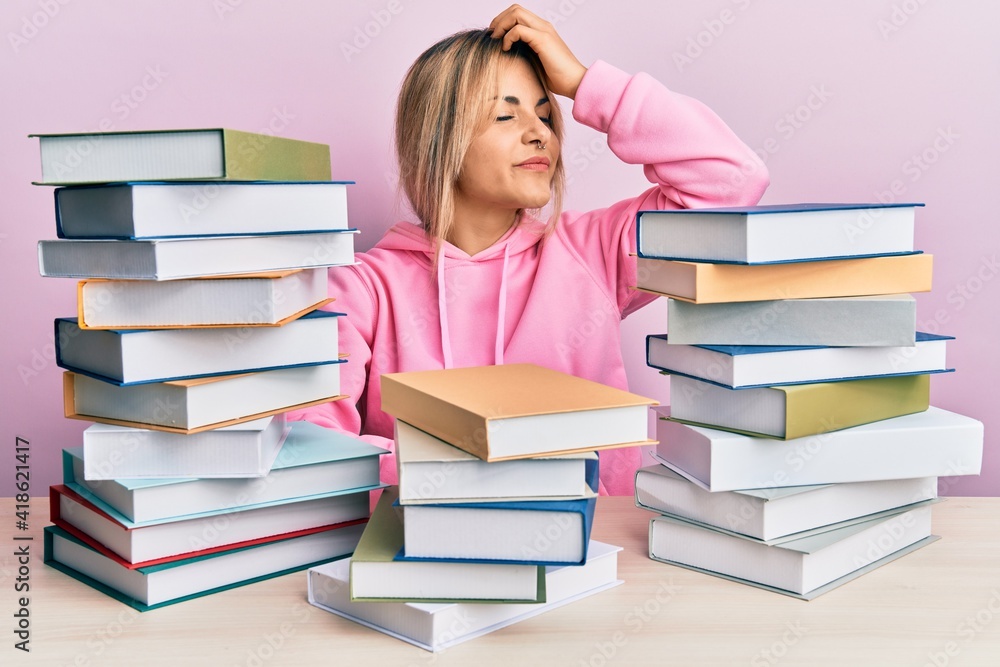 Young caucasian woman sitting on the table with books smiling confident touching hair with hand up gesture, posing attractive and fashionable Young caucasian woman sitting on the table with books smiling confident touching hair with hand up gesture, posing attractive and fashionable