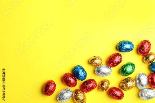 Chocolate eggs wrapped in colorful foil on yellow background, flat lay. Space for text