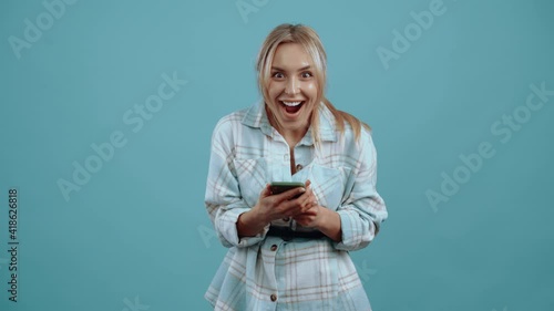 The young woman who uses the phone, amazed looks at the camera and smiles, wide smile, beautiful, emotional Beautiful mature young woman, blonde, in a shirt, blue. Isolated on blue background, Concept