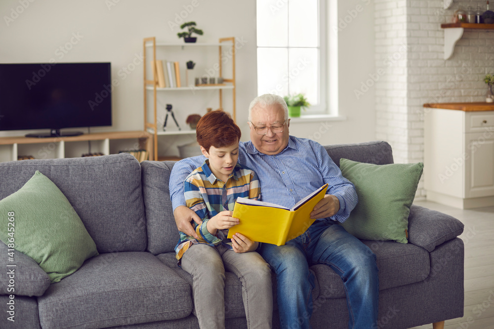 Senior family members and children spend time together: Happy grandfather reading book of good stories to his teen grandson while sitting on comfortable sofa in cozy living-room