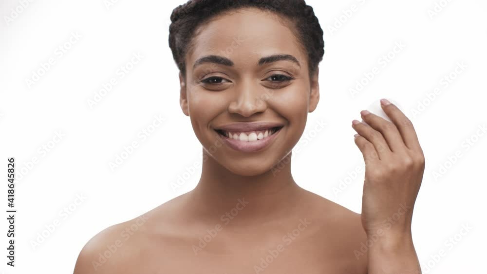 Make up removal. Young pretty black lady cleaning her face with cotton pad, moisturizing her skin, smiling to camera Make up removal. Young pretty black lady cleaning her face with cotton pad, moisturizing her skin, smiling to camera