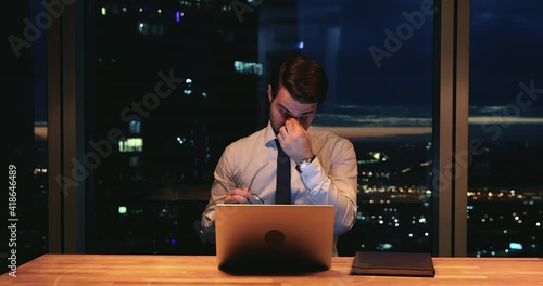 Stressed young caucasian businessman taking off eyeglasses, having painful feelings or dry eyes syndrome due to computer overwork, working overtime alone late in modern office, workaholic concept. photo