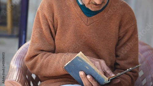 Close up Portrait of old Indian man reading book while sitting on chair at home. Leisure activity of retired people. Senior people lifestyle