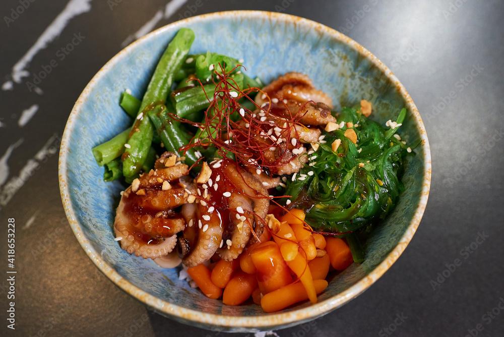 A top view of a poke bowl with octopuses, Chuka Wakame seaweed, carrot, peanut, sesame, white rice on a black stone table. A top view of a poke bowl with octopuses, Chuka Wakame seaweed, carrot, peanut, sesame, white rice on a black stone table.