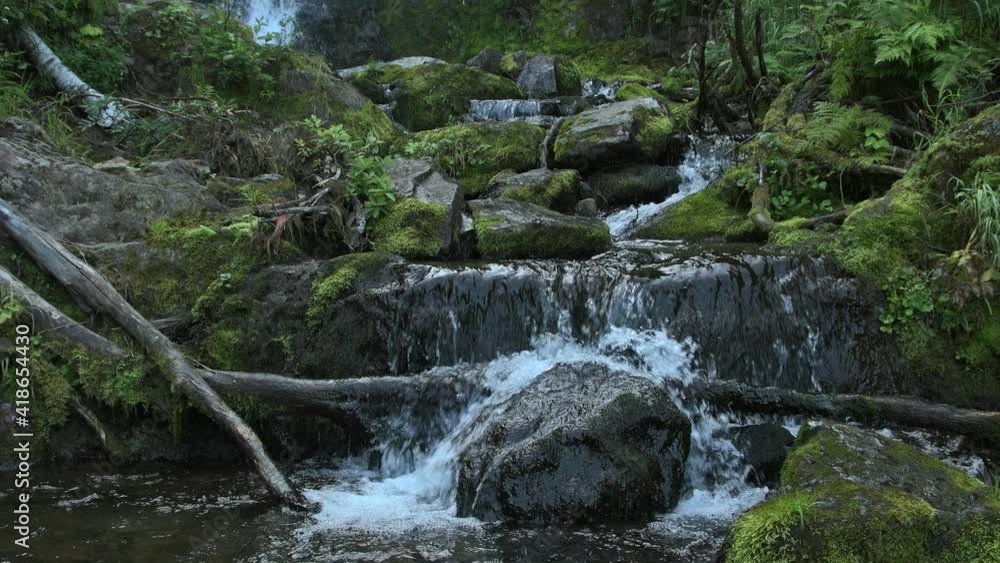 Panorama of mountain waterfall in tropical forest. Rocks along stream are covered with moss and green grass. Old tree trunks lie on shore. Creek foams and flows along cliffs Panorama of mountain waterfall in tropical forest. Rocks along stream are covered with moss and green grass. Old tree trunks lie on shore. Creek foams and flows along cliffs