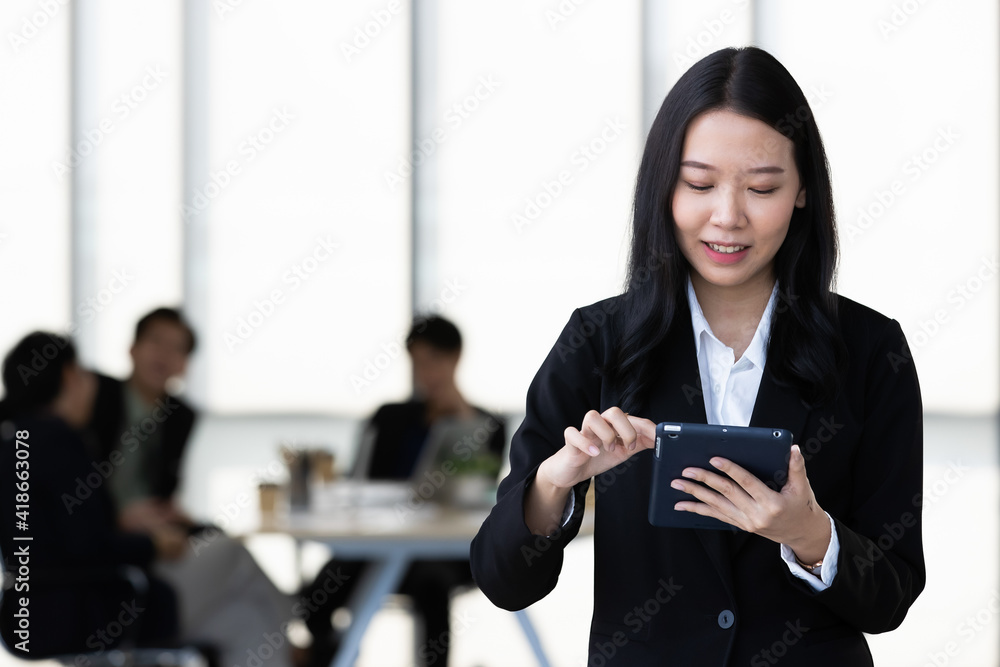 Young Asian businesswoman in black suit standing and holding tablet computer posing to camera in office with her team talking in blur background Young Asian businesswoman in black suit standing and holding tablet computer posing to camera in office with her team talking in blur background