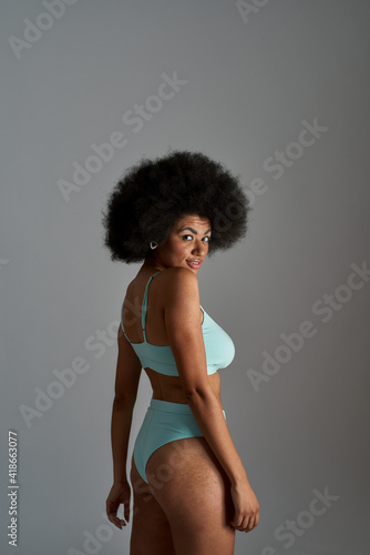 Attractive young african american woman with afro hairstyle wearing blue underwear looking playful at camera while posing isolated over grey background