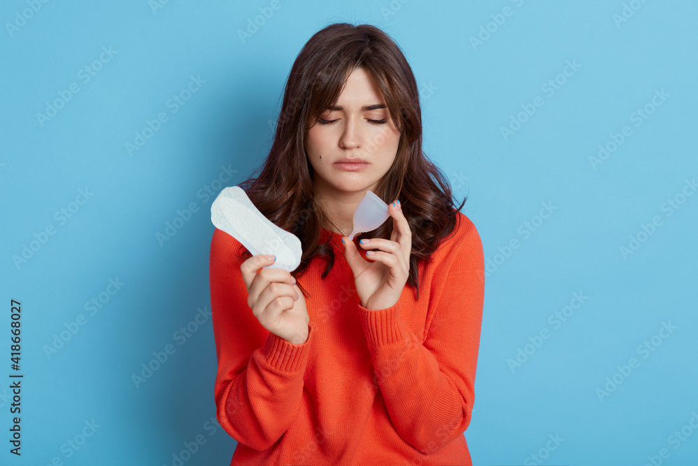 Young beautiful woman European young woman wearing orange sweater making choice between menstrual cup and hygiene pad, suffering from pain, need to choose best hygienic product, isolated on blue wall. Young beautiful woman European young woman wearing orange sweater making choice between menstrual cup and hygiene pad, suffering from pain, need to choose best hygienic product, isolated on blue wall.