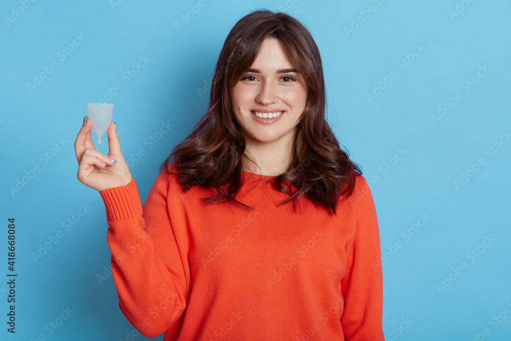 Attractive dark haired female holding in hand plastic menstrual cup isolated over blue background, smiling woman thinks about her personal hygiene product during period, wearing casual orange jumper. Attractive dark haired female holding in hand plastic menstrual cup isolated over blue background, smiling woman thinks about her personal hygiene product during period, wearing casual orange jumper.