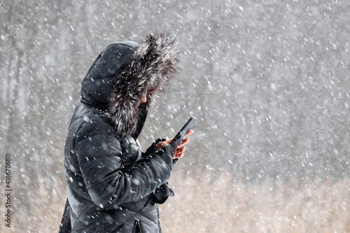 Cold weather and blizzard in city park. Woman in fur hood standing with smartphone in hands during snowfall at march