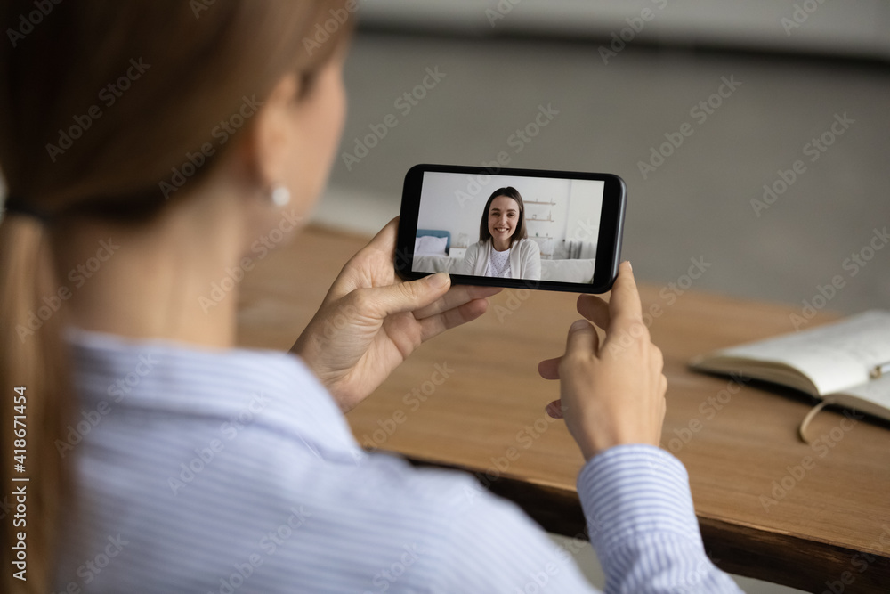 Over shoulder view of female hold cellphone have webcam online conversation with friend or coworker. Woman speak talk on video call on smartphone at home. Communication, virtual event concept.