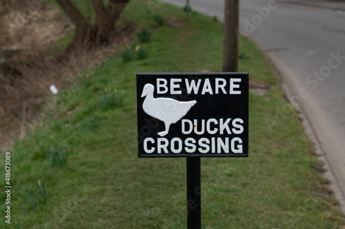 Beware duck crossing sign on some grass, on a river bank. 