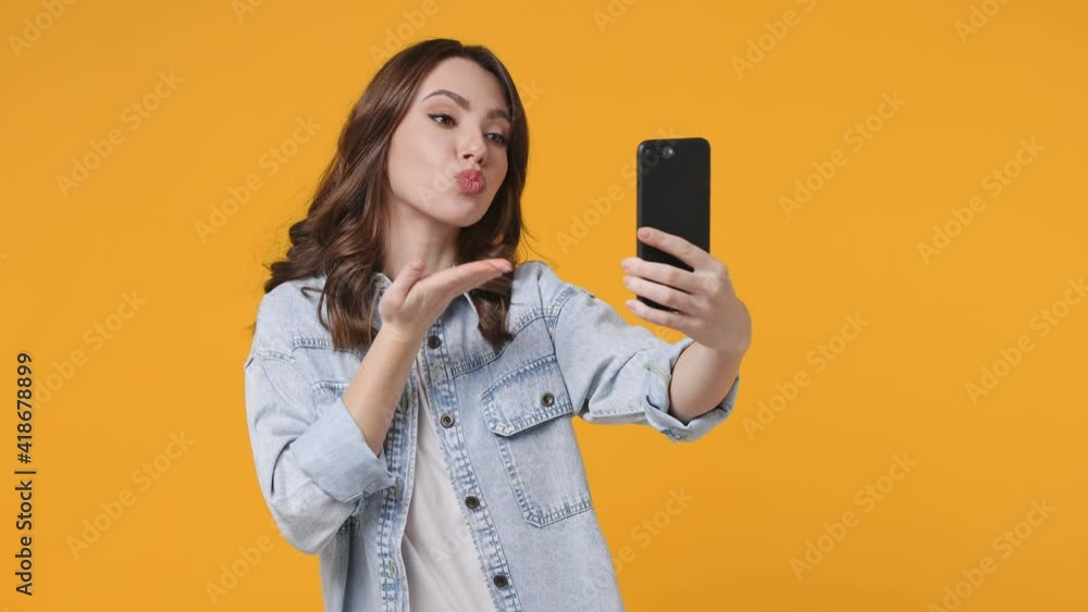 Pretty young woman 20s in denim jacket posing isolated on yellow background studio. People lifestyle concept. Doing selfie shot on mobile phone showing victory sign blinking blowing sending air kiss Pretty young woman 20s in denim jacket posing isolated on yellow background studio. People lifestyle concept. Doing selfie shot on mobile phone showing victory sign blinking blowing sending air kiss