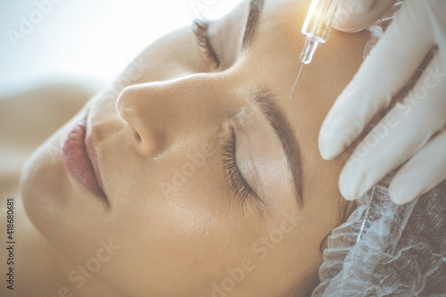 Beautician doing beauty procedure with syringe to face of young brunette woman in sunny clinic. Cosmetic medicine and surgery, beauty injections