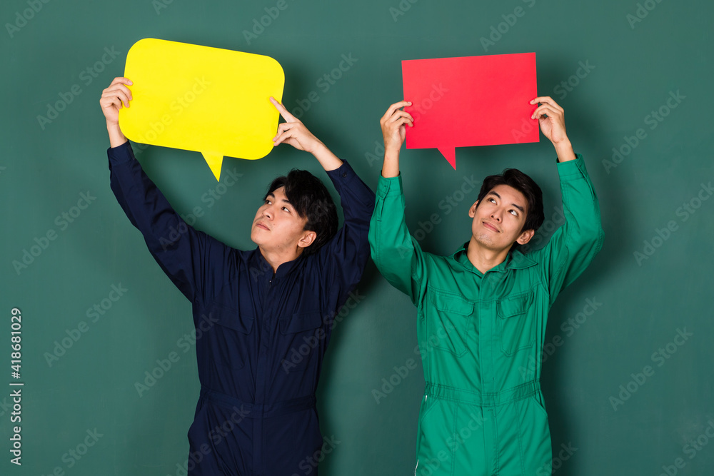 © Bangkok Click Studio - portrait of two young confident handsome Asian men wearing colorful mechanic jumpsuits standing smiling holding a yellow and red text speech bubble over green background