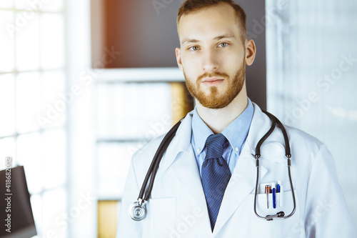 Red-bearded doctor standing straight in sunny clinic near his working place. Portrait of physician. Medicine concept