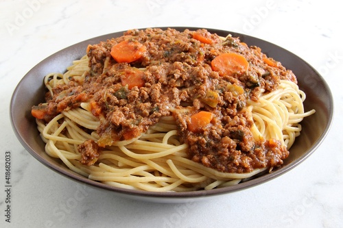 Spaghetti pasta with minced meat, tomato sauce and sliced carrot on a brown ceramic plate on a white marble table. Closeup, selective focus