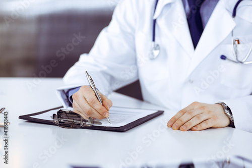 Unknown male doctor sitting and working with clipboard of medication history record in clinic at his working place, close-up. Young physician at work. Perfect medical service, medicine concept