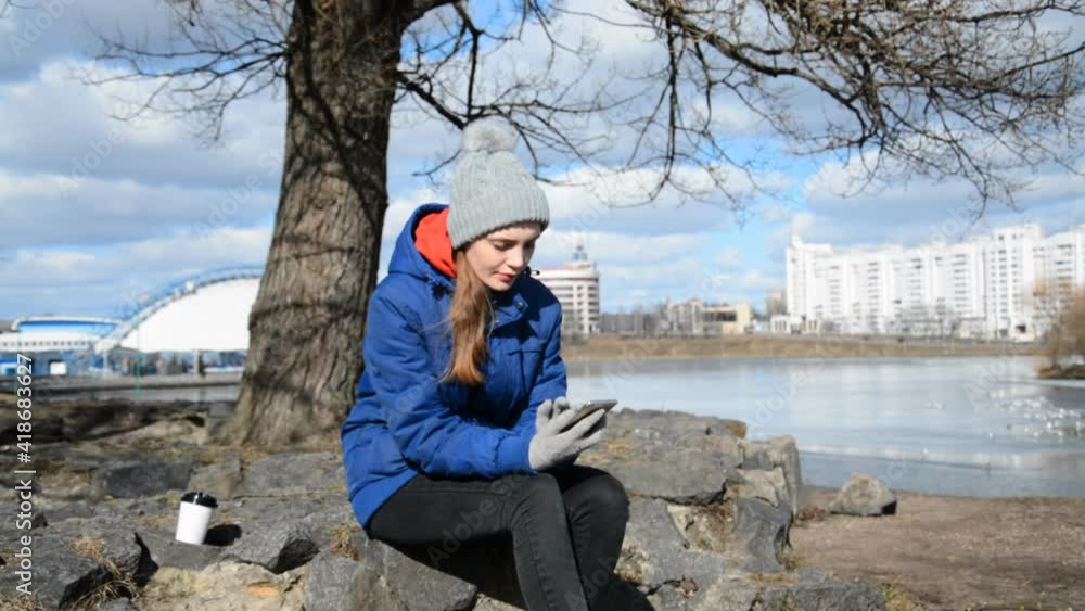 A lonely girl in a blue jacket in spring or autumn on a sunny day sits in a city park and writes a message on a smartphone