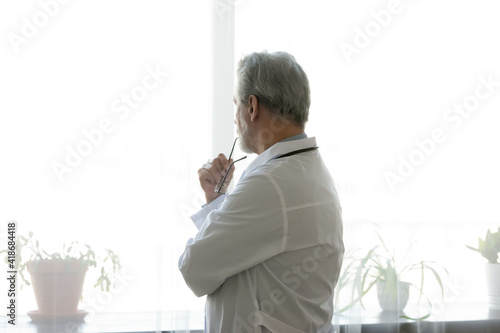 Back view of pensive middle-aged Caucasian male doctor in white medical uniform look in window distance thinking pondering. Thoughtful mature man GP make decision about patient treatment in clinic.