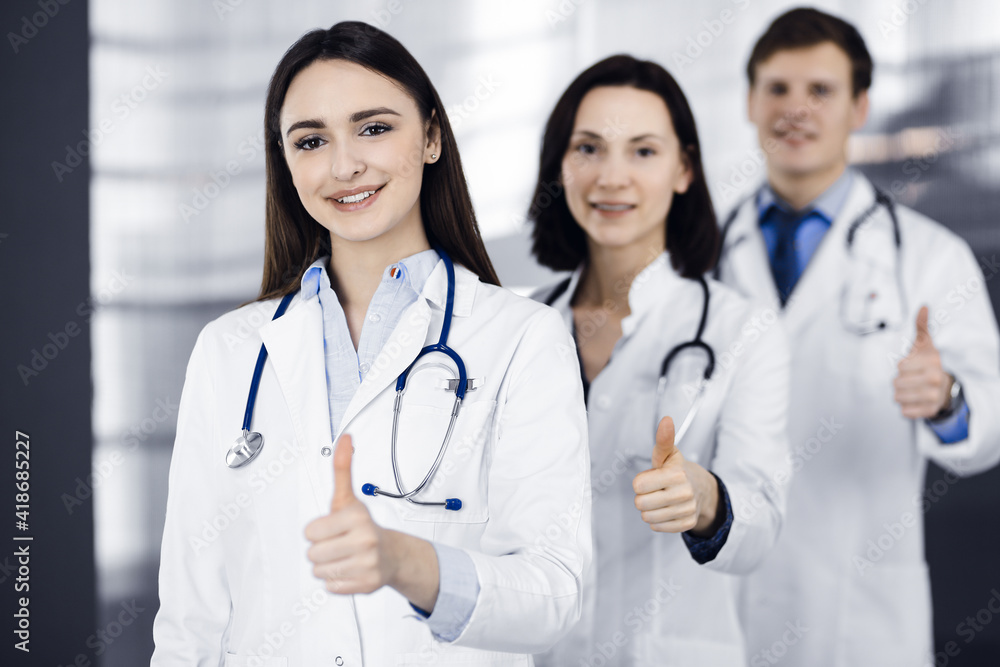Group of young cheerful doctors is standing as a team with thumbs up in a hospital office and is ready to help patients. Medical help, insurance in health care, best disease treatment and medicine Group of young cheerful doctors is standing as a team with thumbs up in a hospital office and is ready to help patients. Medical help, insurance in health care, best disease treatment and medicine