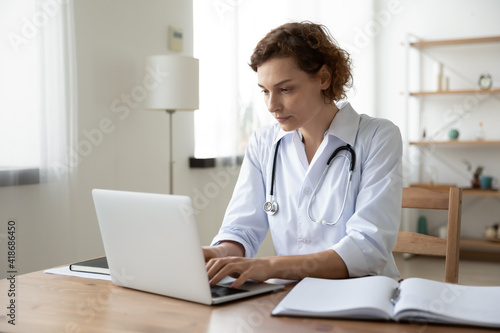 Focused young Caucasian woman doctor in white medical uniform sit at desk in hospital work on laptop online. Concentrated female nurse or GP look at computer screen consult patient on gadget.
