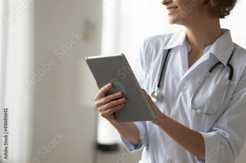 Close up of smiling Caucasian female doctor in white medical uniform use modern tablet gadget in clinic. Happy woman nurse or GP consult patient on pad device in modern hospital. Technology concept.