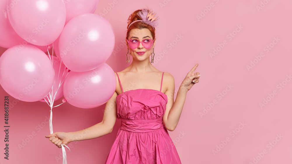 Dreamy fashionable woman wears sunglasses and festive dress points away on blank space holds bunch of inflated balloons enjoys holiday isolated over pink background. Special occasion concept