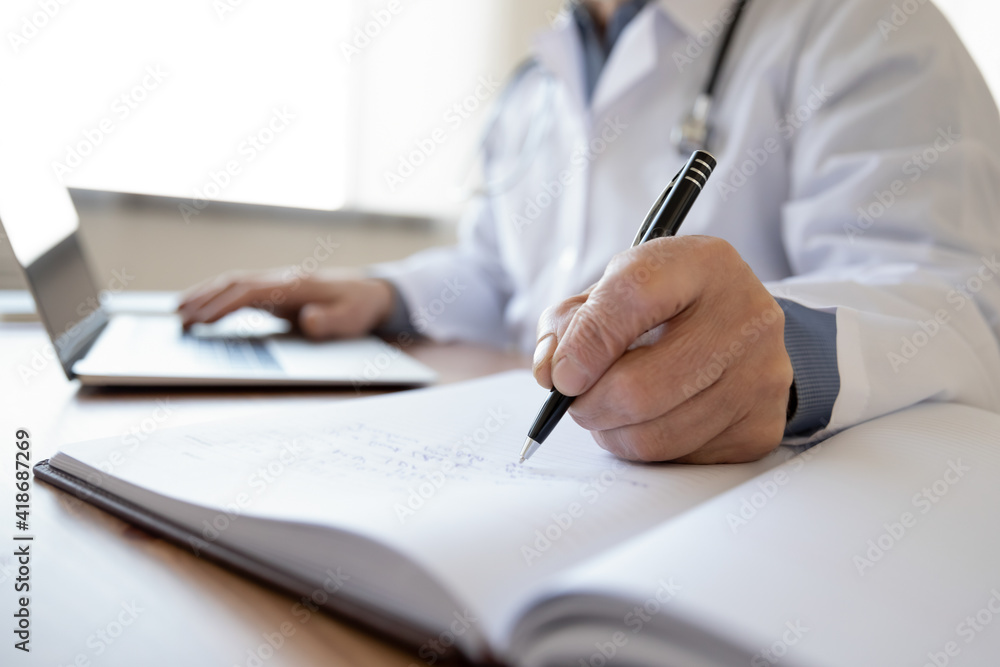 Crop close up of middle-aged male doctor write in medical journal fill patient information use laptop online. Mature man GP or therapist make notes work distant on computer in clinic or hospital. Crop close up of middle-aged male doctor write in medical journal fill patient information use laptop online. Mature man GP or therapist make notes work distant on computer in clinic or hospital.