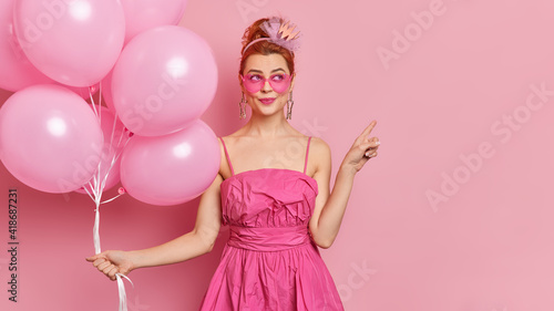 Dreamy fashionable woman wears sunglasses and festive dress points away on blank space holds bunch of inflated balloons enjoys holiday isolated over pink background. Special occasion concept
