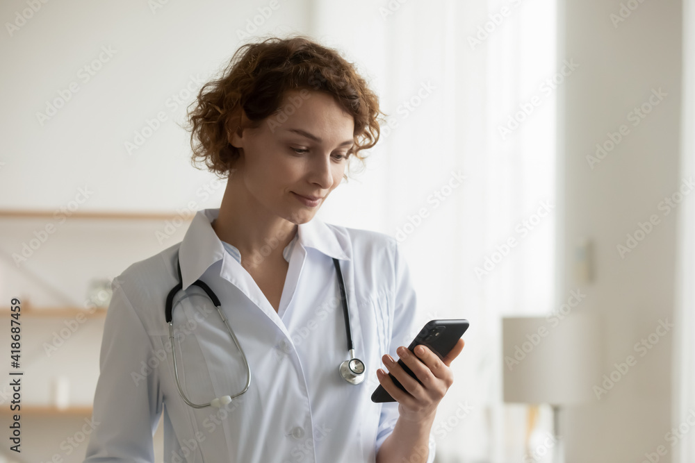 Happy young Caucasian female nurse or GP in white uniform look at cellphone screen use gadget in hospital. Smiling woman doctor consult patient or talk with clinic client on video call on smartphone. Happy young Caucasian female nurse or GP in white uniform look at cellphone screen use gadget in hospital. Smiling woman doctor consult patient or talk with clinic client on video call on smartphone.