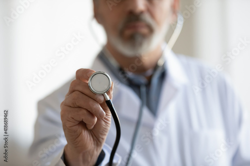 Crop close up of mature male doctor in white medical uniform hold stethoscope examine patient in clinic. Elderly man GP or therapist use phonendoscope do checkup in hospital. Healthcare concept.