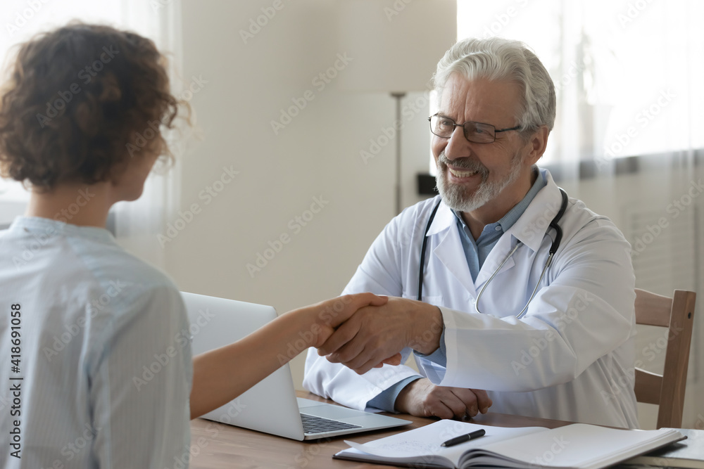 Obraz premium Smiling elderly male doctor shake hang greet get acquainted with female patient at meeting in clinic. Happy mature man GP handshake woman close health insurance deal at consultation in hospital.