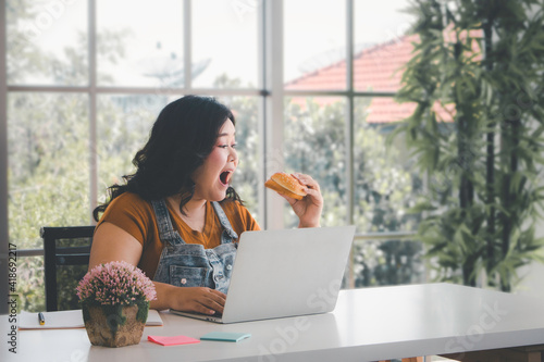 Asian plus size woman with computre laptop eating hamburger working from home, Working lifestyle concept photo