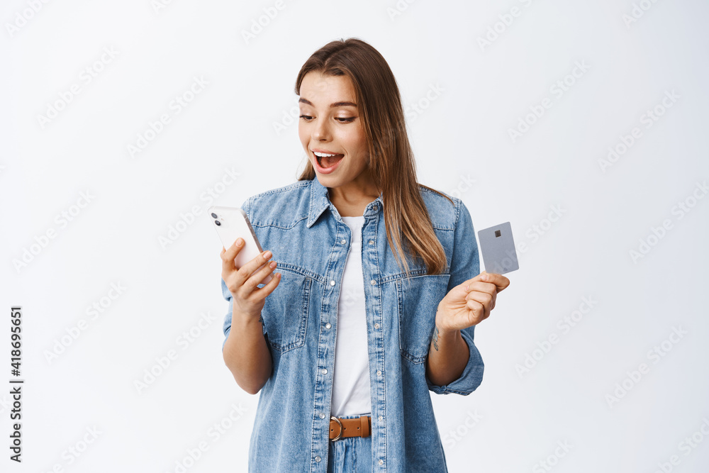 Online shopping. Portrait of excited young woman reading smartphone screen and holding plastic credit card, paying for internet purchase, making an order, white background Online shopping. Portrait of excited young woman reading smartphone screen and holding plastic credit card, paying for internet purchase, making an order, white background